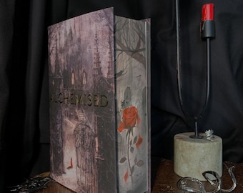 May include: A hardcover book with the title "ALCHEMISED" in gold lettering. The cover features a gothic illustration of a gate and a building, with a red rose on the spine. A black candle holder with red candles is to the right.