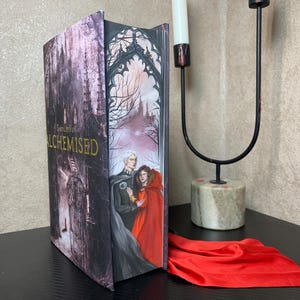 Könnte beinhalten: Ein Hardcover-Buch mit dem Titel "ALCHEMISED" mit einer gotischen Illustration eines Schlosses und Figuren in einem roten Umhang. Ein schwarzer Metallleuchter mit zwei weißen Kerzen und ein rotes Tuch sind ebenfalls vorhanden.