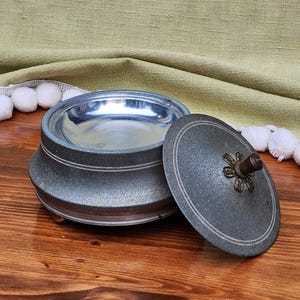 Puede incluir: Una caja de metal vintage de color plateado con un diseño floral en la tapa. La caja tiene un interior brillante y reflectante y se apoya en tres pequeños pies.
