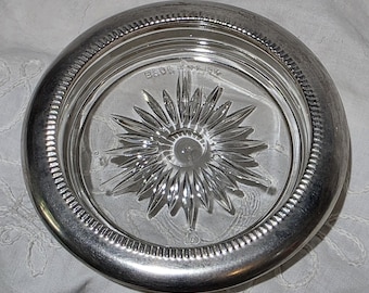 May include: A round glass coaster featuring a clear center with a pressed starburst pattern and a metallic silver-toned rim with a textured, ribbed edge. The word MADE IN USA is embossed in small letters near the top of the glass center.