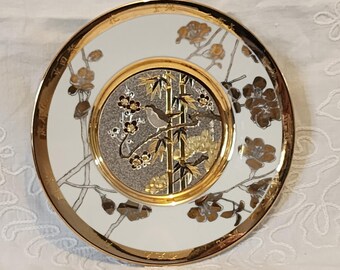 Könnte beinhalten: Ein weißer Porzellanteller mit goldfarbenem Rand und einem silbernen und goldenen Design eines Vogels, der auf Bambuszweigen sitzt. Der Teller hat einen floralen Rand.