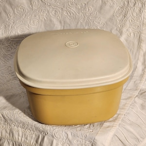 Puede incluir: Un recipiente Tupperware vintage con una base de color amarillo mostaza y una tapa color crema. La tapa tiene una hendidura circular en el centro. El recipiente es de forma cuadrada y está sobre una superficie blanca estampada.