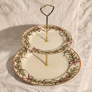 Puede incluir: Bandeja de dos niveles con asa dorada. Los platos blanquecinos están decorados con un diseño festivo verde y rojo. Los bordes están adornados con dorado. Ideal para servir postres o aperitivos.