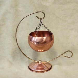 May include: A copper-colored bowl suspended by a chain from a curved metal stand. The bowl has a rounded shape and reflects light. The stand is made of a lighter-colored metal and has a circular base. The overall design is decorative.