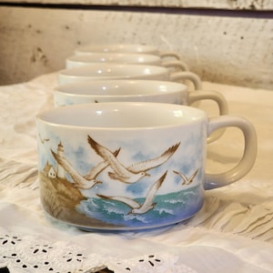 May include: A set of six white ceramic mugs with a coastal design. Each mug features a lighthouse, seagulls, and ocean waves in shades of blue, brown, and white. The mugs have a rounded shape and a light gray handle.