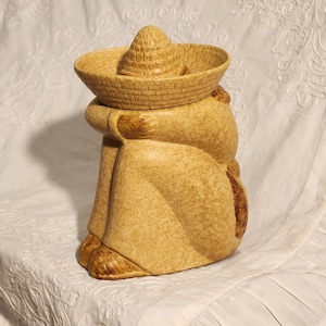 May include: A tan ceramic cookie jar in the shape of a person wearing a sombrero. The jar has a textured surface and is in a seated position. The sombrero lid is removable, and the jar has brown accents.