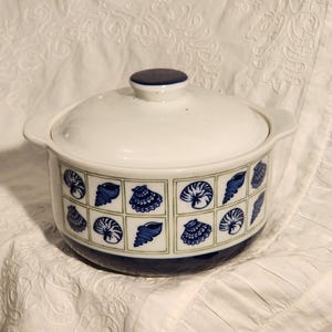 May include: White ceramic casserole dish with a lid. The dish features a blue and white seashell pattern around the sides and a dark blue rim. The lid has a dark blue knob.