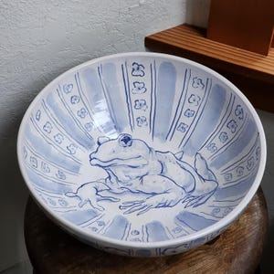 May include: A white ceramic bowl with a blue and white design featuring a frog. The bowl has a striped pattern with small flower designs.