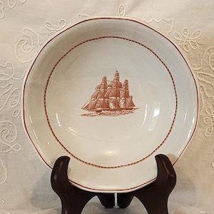 Puede incluir: Cuenco de cerámica blanco con una ilustración marrón de un barco en el centro. El cuenco tiene un borde marrón en forma de cuerda y un borde marrón. Se muestra sobre un soporte de madera marrón oscuro.