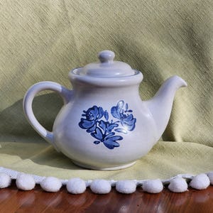 May include: A white teapot with a blue floral design. The teapot has a round body and a spout on the side. The teapot is sitting on a white and green tablecloth.