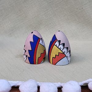 May include: A pair of ceramic salt and pepper shakers with a pink base and colourful geometric designs. The shakers are egg-shaped and feature blue, yellow, red, and white accents.