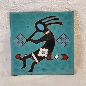 May include: Square turquoise tile featuring a Kokopelli figure in black, white, red, and blue. The Kokopelli is playing a flute. The tile has a speckled texture and a brown border. Decorative tile.