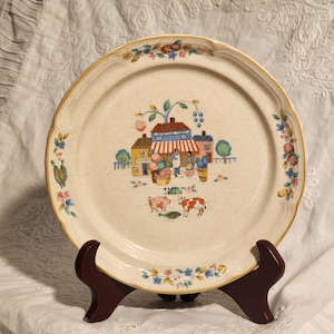 May include: A cream-colored ceramic plate with a village scene in the center. The scene includes buildings, people, and animals. The rim is decorated with floral and fruit motifs, and a gold trim. The plate is displayed on a brown wooden stand.