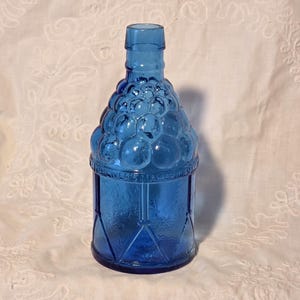 Puede incluir: Una botella de vidrio azul vintage con un diseño único de racimo de uvas en la parte superior. La botella tiene una base rectangular con patrones geométricos y el texto "WELLERS AMERICAN" es visible. La botella es de vidrio azul transparente.