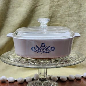 May include: A vintage, pale pink casserole dish with a clear glass lid and handle. The dish has a blue floral design. It is displayed on a clear glass cake stand.