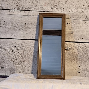 May include: A rectangular, vintage-style mirror with a narrow, golden-brown frame. The mirror is tall and slender, reflecting a light-colored interior. It rests on a white, textured surface against a weathered, wooden backdrop.