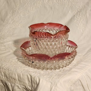 以下が含まれることがあります： ルビーレッドの縁取りが施された透明なガラスのボウルと、お揃いの皿。ボウルと皿にはダイヤモンド模様があります。ボウルと皿の縁はスカラップエッジです。ボウルは皿の上にあります。