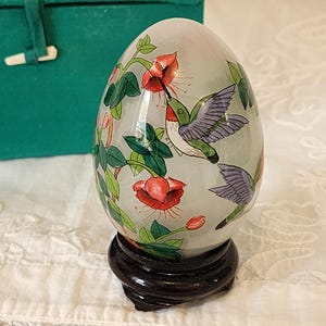 May include: An ornamental egg featuring a painted design of hummingbirds and red flowers. The egg is displayed on a dark wooden stand. A green velvet box is in the background.