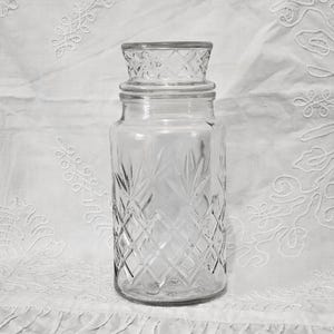 Op de afbeelding: Een glazen pot met een transparante kleur en een ruitvormig patroon en een deksel. De pot is leeg en heeft een vintage uitstraling.