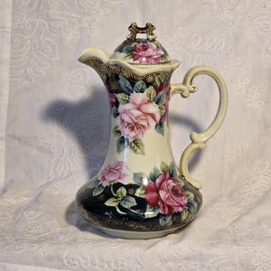 Può includere: Una brocca vintage in porcellana con coperchio, con un corpo color crema ornato di rose rosa e foglie verdi. La base e il bordo superiore presentano un motivo blu scuro e oro. Il manico e il beccuccio sono color crema con dettagli dorati.