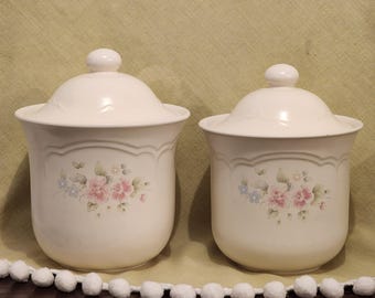 Könnte beinhalten: Zwei weiße Keramikdosen mit Blumendesigns. Die Dosen haben Deckel und sind mit rosa und blauen Blumen verziert.