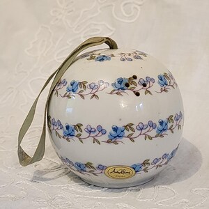 May include: A white, spherical ornament with blue floral patterns and a beige ribbon for hanging. The ornament has a label that reads "Andre Richard Japan". The floral design features blue roses and green leaves.