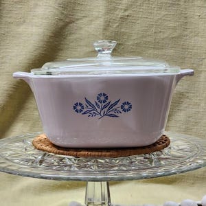 May include: White casserole dish with a glass lid and a blue floral design. The dish is sitting on a glass cake stand.