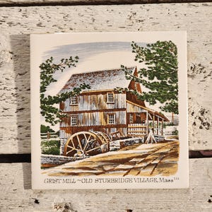May include: Square ceramic tile featuring a detailed illustration of the Grist Mill in Old Sturbridge Village, Massachusetts. The artwork depicts a wooden mill with a water wheel, surrounded by trees and a road. The tile has a white background.