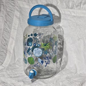 Può includere: Un distributore di bevande in vetro trasparente con coperchio e manico blu e un rubinetto blu. Il distributore è decorato con disegni di conchiglie e coralli blu, verdi e bianchi. Il distributore è alto circa 25 cm.