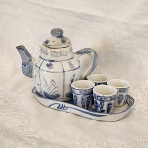 May include: A blue and white ceramic tea set, including a teapot, four small cups, and a serving tray. The teapot and cups have a blue floral and geometric pattern. The set is presented on a white, textured surface.