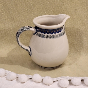 Może przedstawiać: Biały dzbanek ceramiczny z niebiesko-zielonym wzorem kwiatowym wokół górnej krawędzi. Dzbanek ma zaokrąglone ciało i uchwyt.