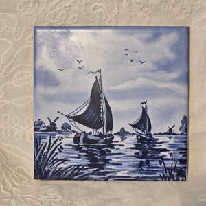 May include: Square ceramic tile with a blue and white nautical scene. The artwork shows sailboats on water, with windmills and birds in the background. The colour scheme is predominantly blue and white.