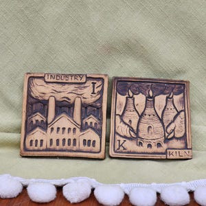 Può includere: Due piastrelle quadrate in ceramica marrone con disegni intagliati. Una piastrella recita "INDUSTRY" e raffigura una fabbrica. L'altra piastrella recita "KILN" e mostra tre forni. Le piastrelle sono su una superficie verde.