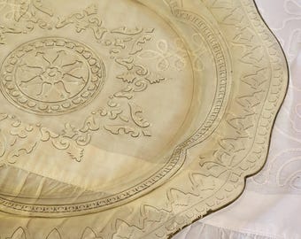 May include: A vintage, pale yellow glass serving tray with an ornate, embossed design. The tray features a scalloped edge and detailed floral and geometric patterns. The tray is resting on a white, ruffled fabric.