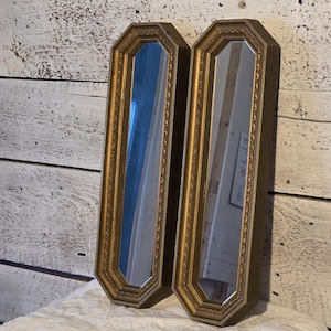 May include: Two vintage, long, rectangular mirrors with gold-colored, ornate frames. The mirrors have an octagonal shape and reflect the surrounding environment. The frames have a detailed, textured design. The mirrors are displayed against a white wooden background.