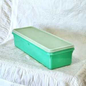 Puede incluir: Un recipiente rectangular de plástico verde vintage con una tapa verde claro. El recipiente está sobre una superficie blanca bordada. El recipiente es una caja de almacenamiento.