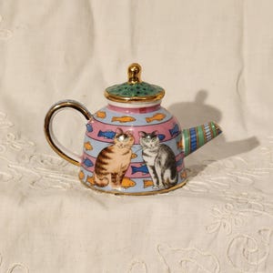 May include: A decorative ceramic teapot featuring two cat illustrations. The teapot has a light blue background with pink stripes and fish designs. The handle and spout are gold-trimmed, and the lid is green with a gold knob.