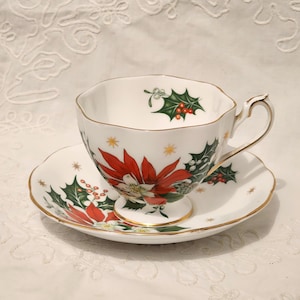May include: A white teacup and saucer set with gold trim. The cup and saucer are decorated with a red poinsettia flower, green holly leaves, and gold stars. The cup handle is gold.