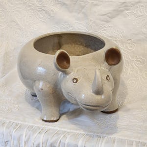 May include: A ceramic planter shaped like a rhinoceros. The planter is white with brown ears and a speckled finish. It has a wide opening at the top for planting.
