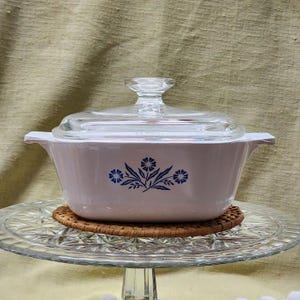 May include: White casserole dish with a glass lid and a blue floral design. The dish is sitting on a clear glass cake stand.