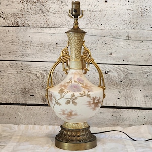 May include: An antique table lamp with a white ceramic body adorned with floral designs in shades of purple and gold. The lamp features brass accents, including a decorative base, handles, and a lamp holder. The lamp is connected to a black power cord.