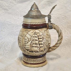 May include: A vintage beer stein with a detailed nautical scene carved into the ceramic body. The stein features a metal hinged lid and handle, with a rope-like design around the top. The base has the text "The Bark & Staysail Stein".