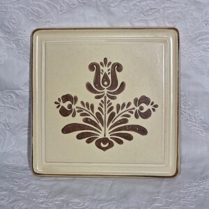 May include: Square ceramic tile with a cream-colored center and a brown floral design. The tile has a brown border and a raised edge. The floral design features stylized flowers and leaves, creating a vintage aesthetic.
