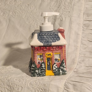 May include: A ceramic soap dispenser shaped like a toy store. The dispenser has a red brick facade, a blue and white snowy roof, and a white pump. The word "TOYS" is displayed on the front. Green trees flank the building.