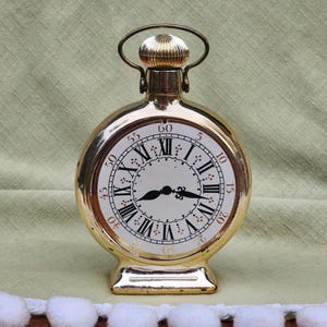 May include: A gold-colored clock-shaped bottle with a Roman numeral clock face and a gold-colored lid. The bottle has a round body and a square base.