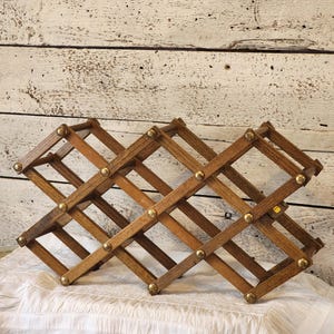 May include: Wooden wine rack with a diamond lattice design. The rack is made of brown wood and features brass-colored rivets at each joint. The rack is designed to hold multiple bottles of wine and is set against a white background.