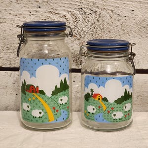 May include: Two glass jars with blue lids and a whimsical farm scene design. The jars feature a red barn, yellow road, green trees, white sheep, and a blue stream.