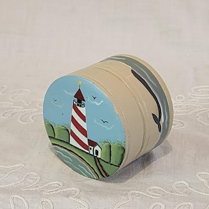 Puede incluir: Caja cilíndrica de madera pintada a mano con un faro a rayas rojas y blancas sobre un fondo azul con pájaros. La caja es de color beige con un diseño de ballena pintada en el lateral.