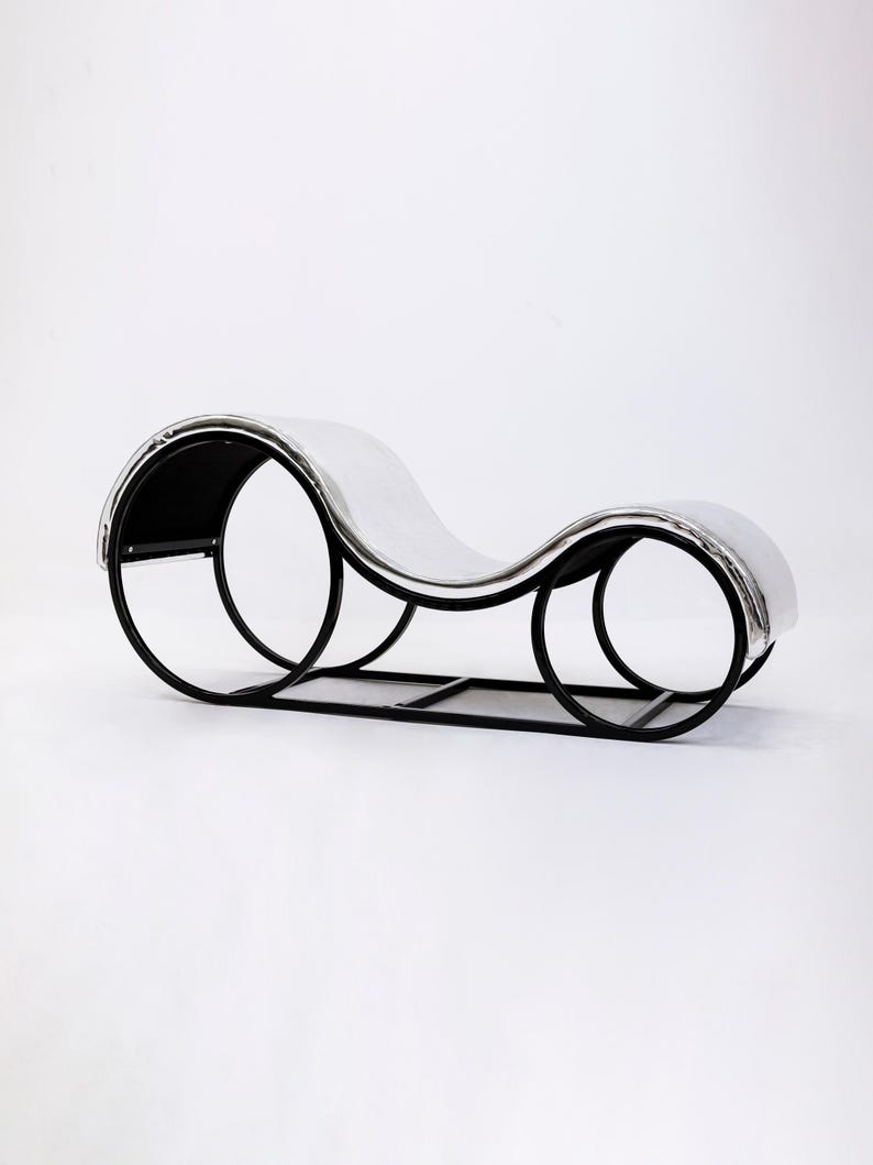 May include: A modern chaise lounge with a curved design. The frame is black metal, supporting a silver-colored, padded seat. The lounge features a unique, open circular design on the sides, creating an elegant and contemporary aesthetic.
