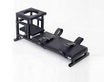 May include: A black wooden bench with a padded surface and adjustable leather straps. It features a raised section with a circular opening and a lower platform. The design is minimalist and functional, with a focus on support and restraint.